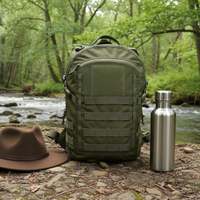 Thumbnail for An olive green curve tactical backpack stands on a rocky riverbank with a forest stream in the background. A brown wide-brim hat rests on the left and a stainless steel water bottle stands on the right.