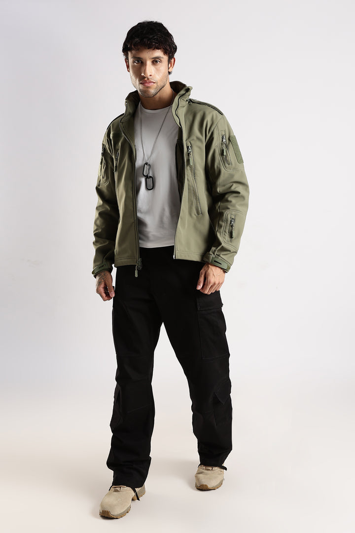 A man modeling an olive green hooded jacket over a light gray t-shirt, black cargo pants, and beige shoes, standing against a plain light gray studio background.