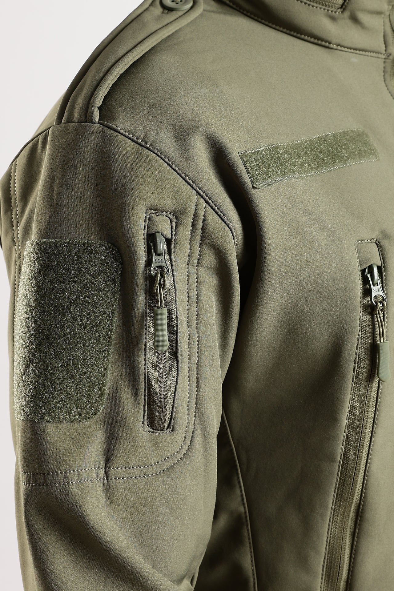 Close-up of an olive green tactical jacket showing the shoulder, sleeve with a zippered pocket and a rectangular velcro patch.