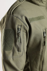Thumbnail for Close-up of an olive green tactical jacket showing the shoulder, sleeve with a zippered pocket and a rectangular velcro patch.